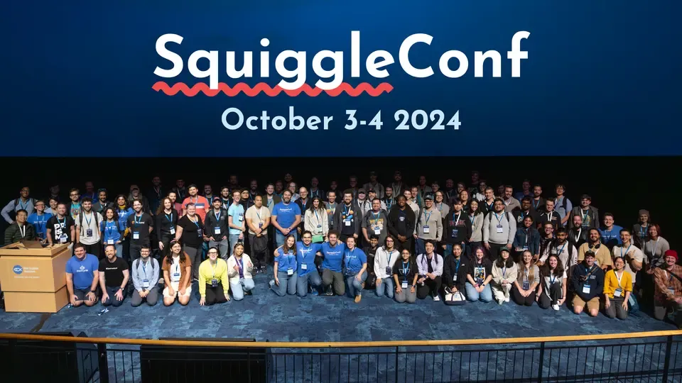 High-level photo of everyone kneeling or standing, smiling, in the front of an auditorium. On the screen, the SquiggleConf October 3-4 2024 logo is visible.